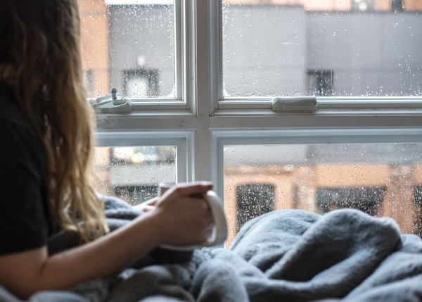 A woman is watching the rain through her windows whilst warming herself with a blanket and mug of hot drink