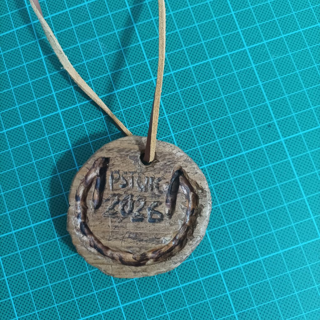A carved wooden medal with the words PSTOIC and the year 2026